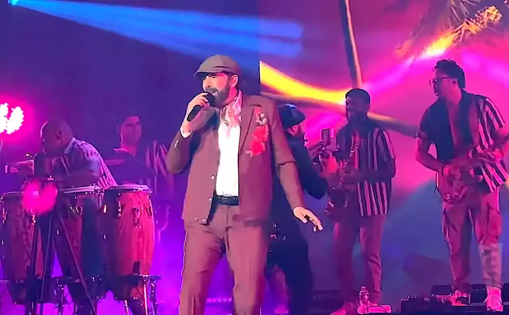 A male singer in a brown suit and cap performs on stage with a band.