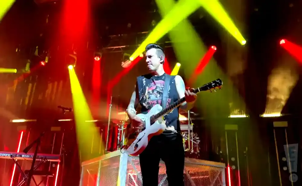 A male guitarist plays a white electric guitar on a stage with red and yellow spotlights.
