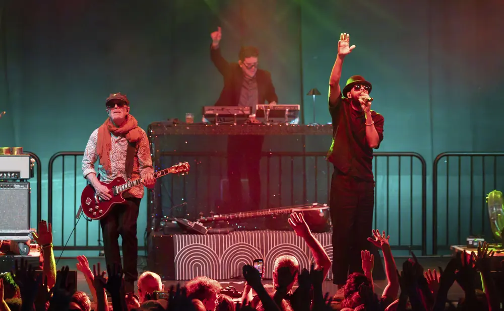 Musicians perform on stage with a crowd cheering in the foreground.