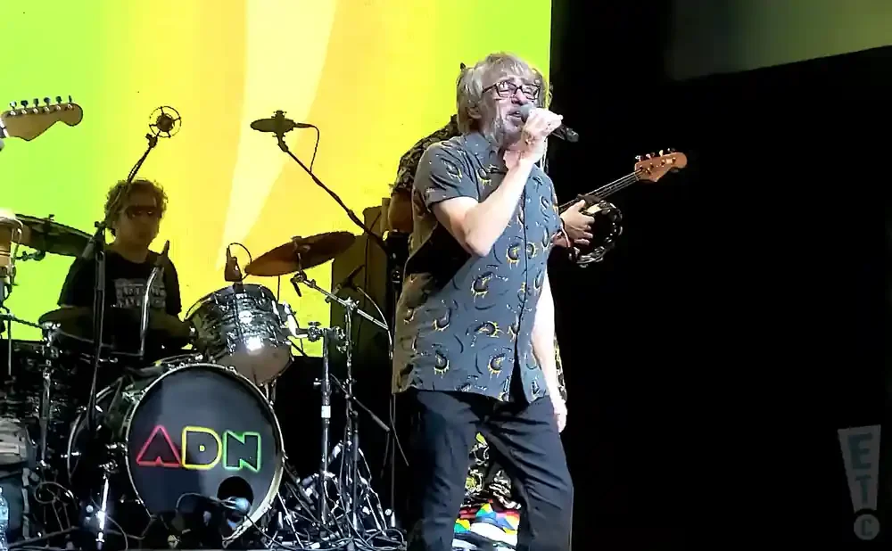 A band performs on stage with a green and yellow backdrop. The lead singer wears a patterned shirt and sings into a microphone.