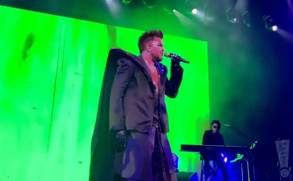 A man in a dark, embellished suit with a cape sings into a microphone on a stage with a bright green backdrop.