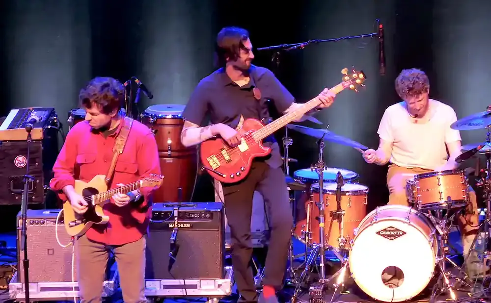 A band plays music on stage, with a guitarist in red, a bassist in a dark shirt, and a drummer in a white shirt.