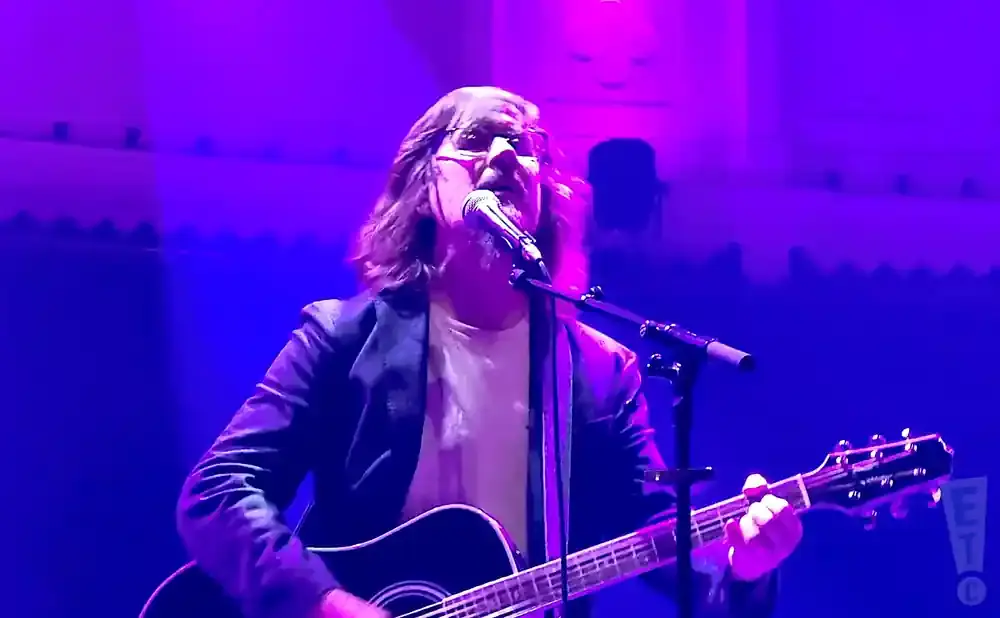 Musician with long hair and glasses plays an acoustic guitar and sings into a microphone on a purple-lit stage.