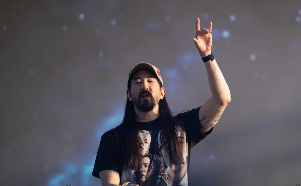 DJ Steve Aoki performs on stage, raising his hand in a rock-on gesture.