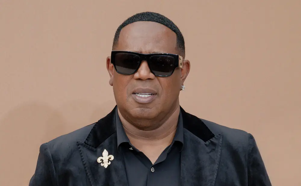Master P wears sunglasses and a black velvet blazer with a fleur-de-lis brooch.