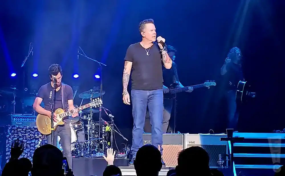 Singer performing on stage with a band, wearing a black t-shirt and jeans, holding a microphone.