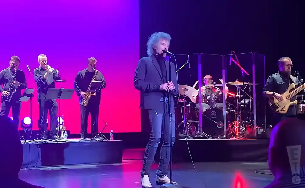 A band performs on stage with a singer in the foreground and horns, drums, and bass guitar players behind him.