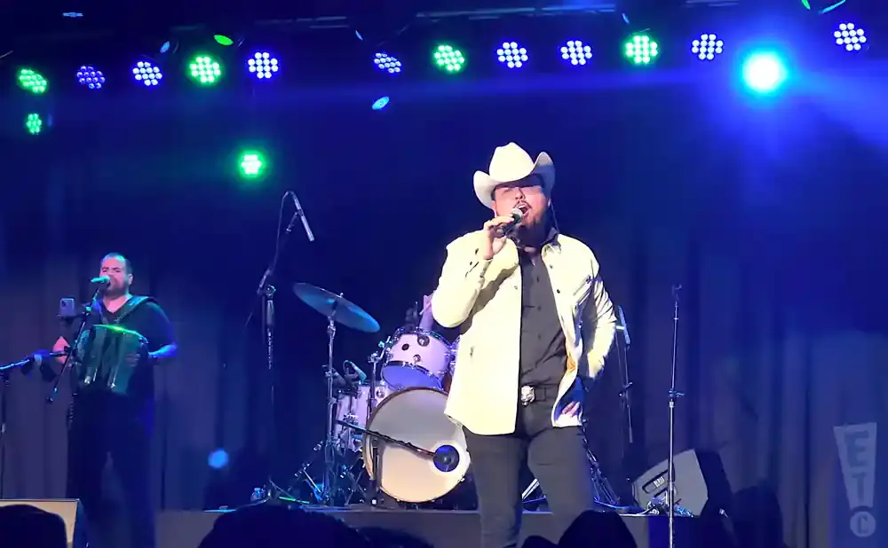 A male singer in a cowboy hat sings into a microphone on stage with a band.