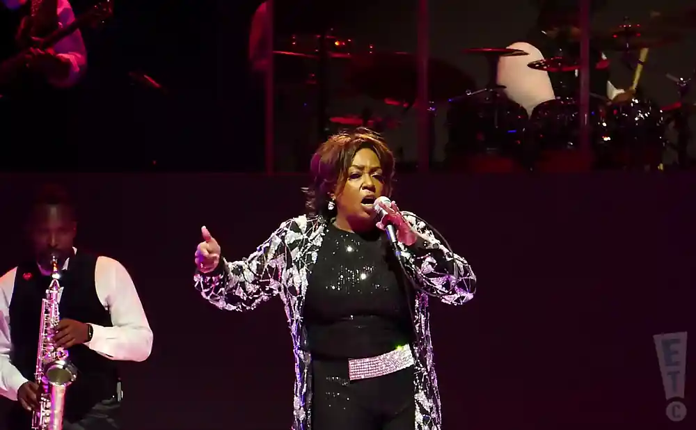 A woman sings into a microphone on stage, wearing a sparkly jacket and pants. A saxophonist plays in the background.