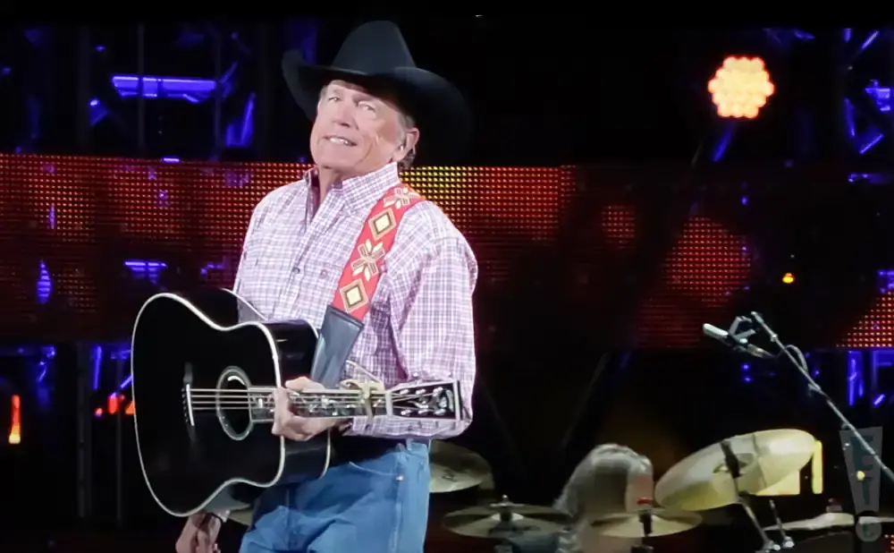 George Strait plays a black acoustic guitar on stage, wearing a cowboy hat and plaid shirt.