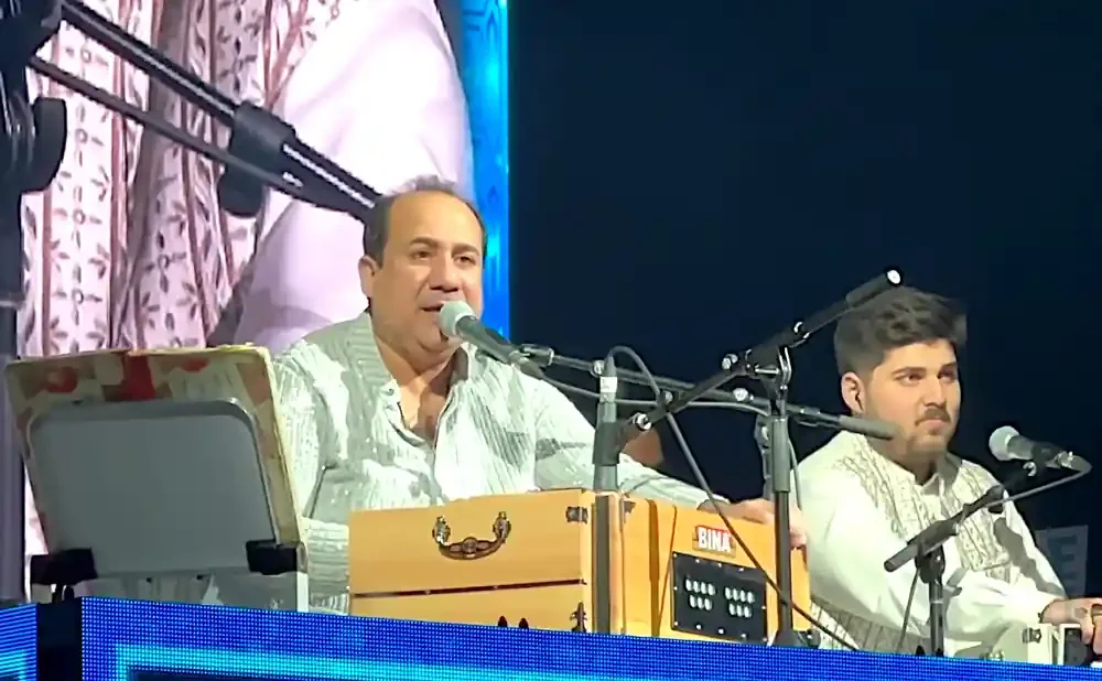 Two men perform music on a stage with microphones and a harmonium.