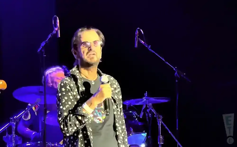 Musician Ringo Starr on stage, holding a microphone and wearing a black shirt with white stars and moons.