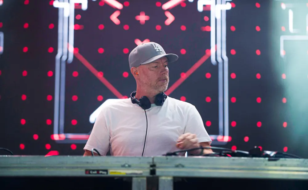 DJ with headphones around neck, wearing a grey cap and white t-shirt, stands behind a DJ console with a blurred stage background.