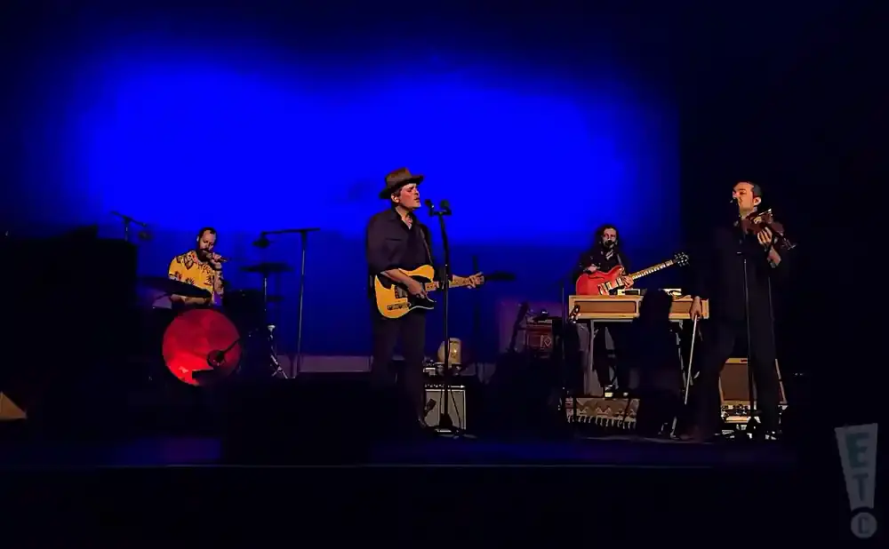 A band performs on a dimly lit stage with a bright blue backdrop. A guitarist sings into a microphone, while a violinist plays.