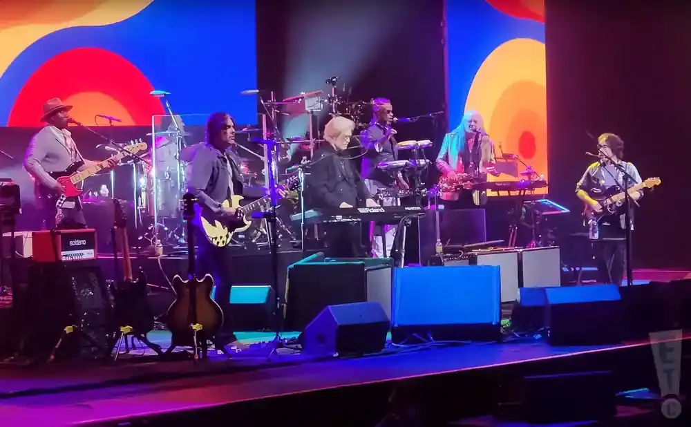 A band performs on stage with colorful, abstract backdrops and bright stage lights.