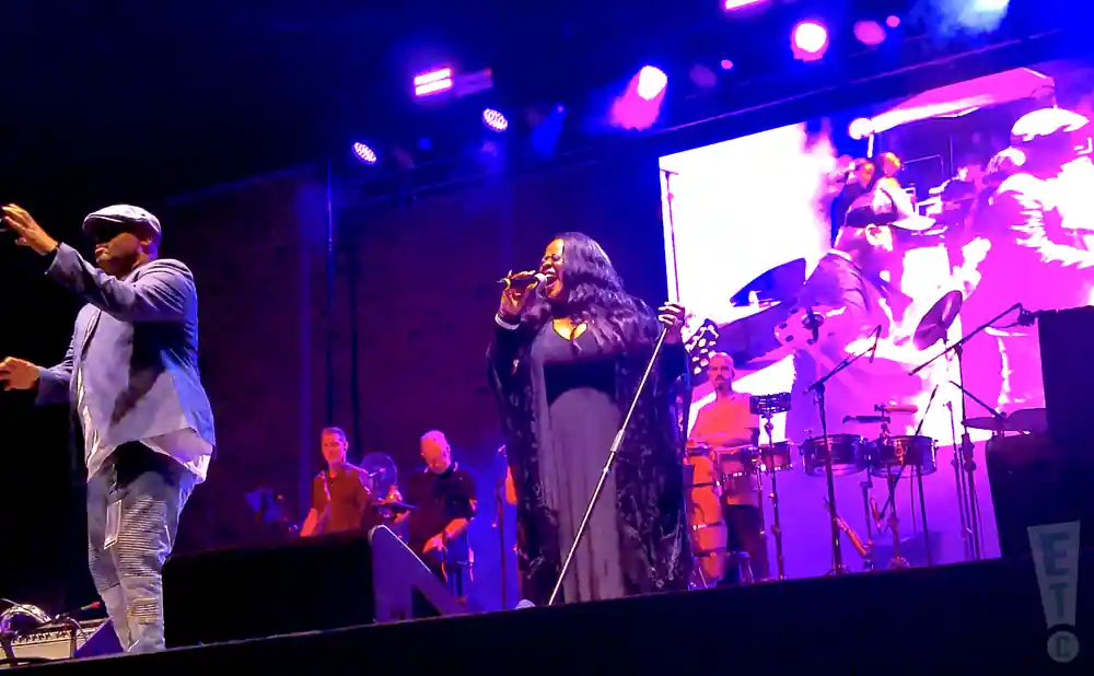 A male singer in a light blue suit and cap gestures to the left as a female singer in a black gown sings into a microphone.