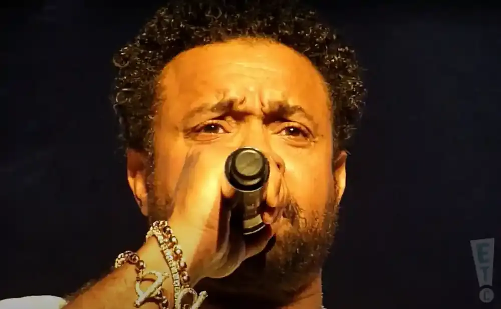 A close-up of a man with curly hair singing into a microphone, wearing gold bracelets with charms.