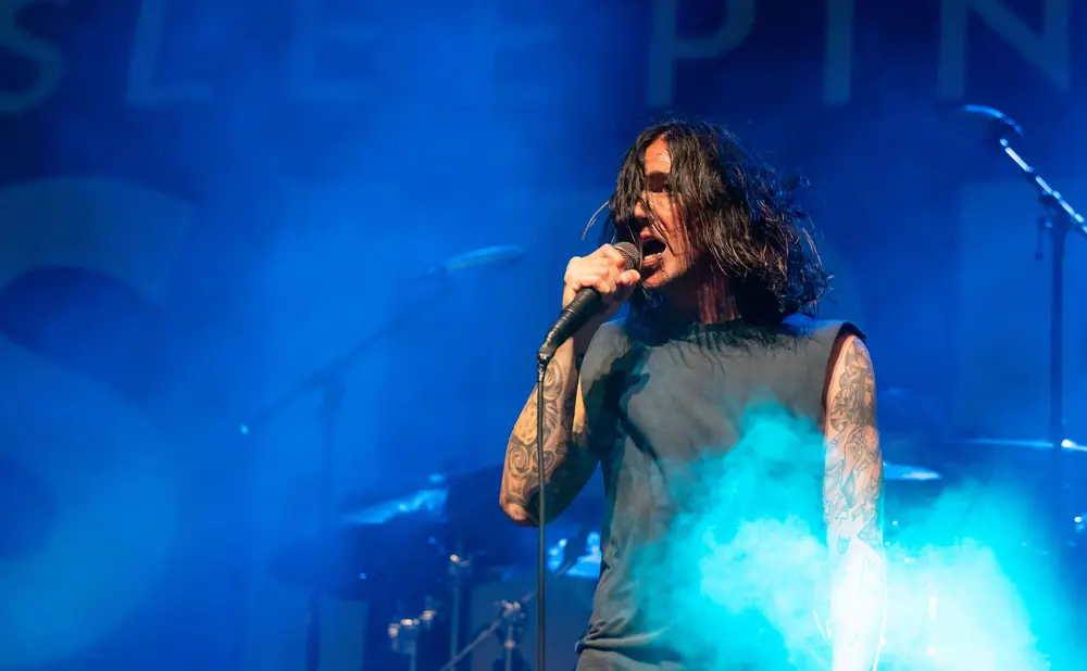 A male singer with long, dark hair sings into a microphone on a stage illuminated by blue lights and smoke.