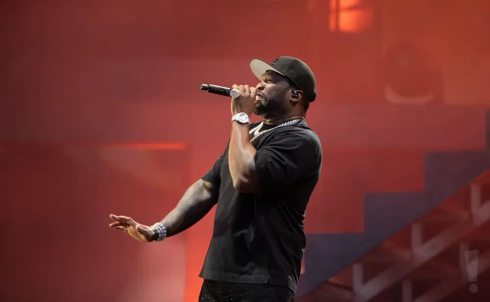 Rapper 50 Cent performs on stage wearing a black t-shirt, black baseball cap, and jeweled pants.