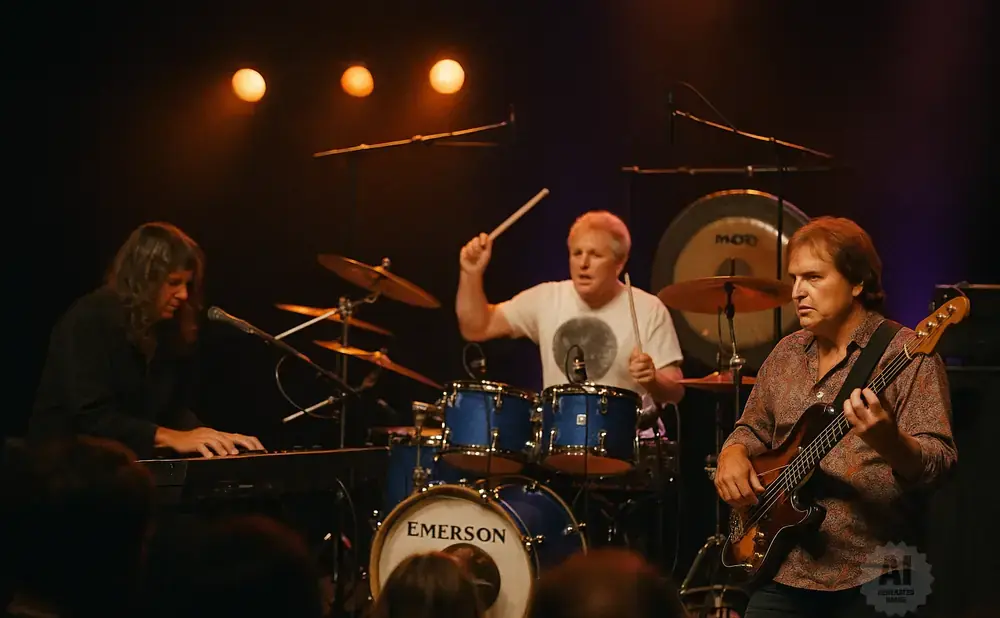 Musicians perform on stage: a keyboardist on the left, a drummer in the center, and a bassist on the right.