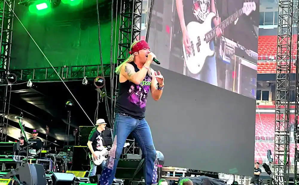 A male singer in a bandana and tank top sings into a microphone on a concert stage with a large screen behind him.