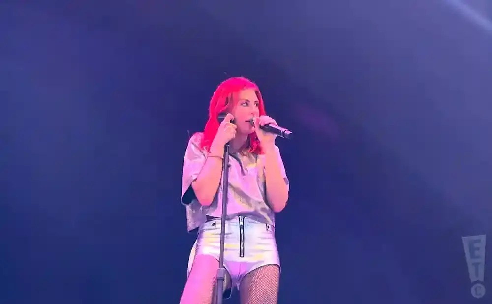 A woman with bright pink hair sings into a microphone on a stage, wearing a silver top and shorts.