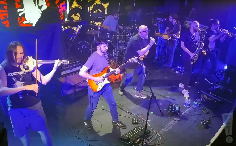 A band performs on stage with a violinist, guitarist, singer, saxophonist, and trumpeter.