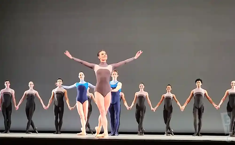A ballet troupe performs on stage, with a central female dancer in a grey leotard with open arms, flanked by male and female dancers in black and blue l…