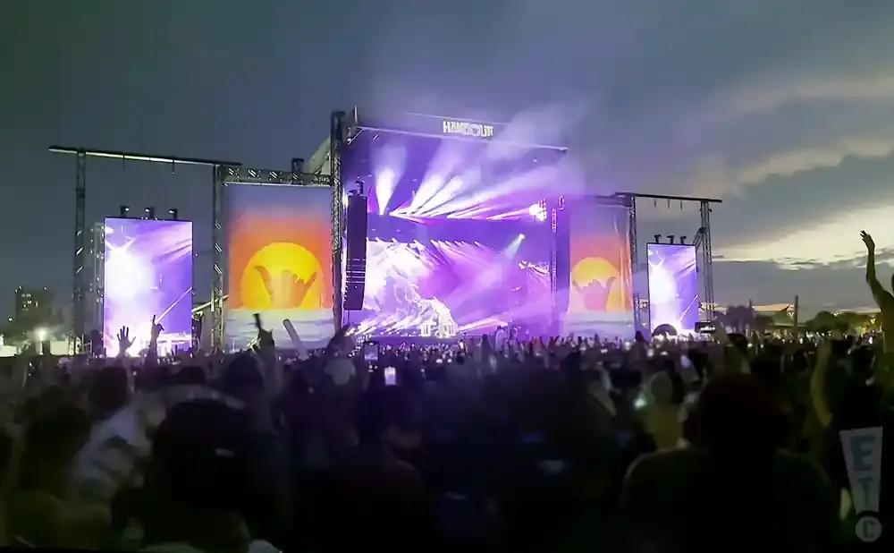 A large crowd watches a concert on a brightly lit stage at dusk.