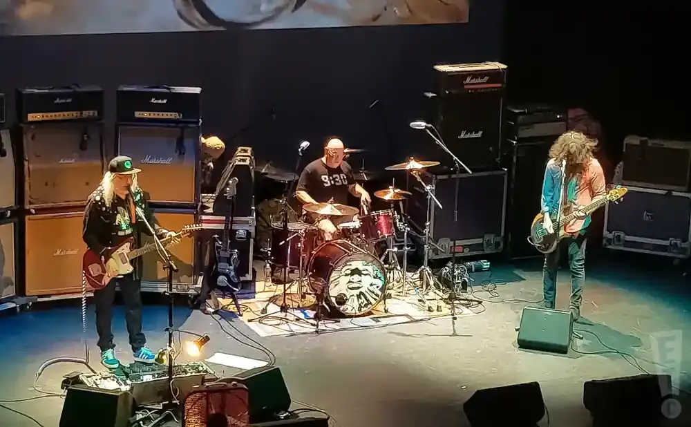 A band performs on stage with large amplifier stacks and a drummer.