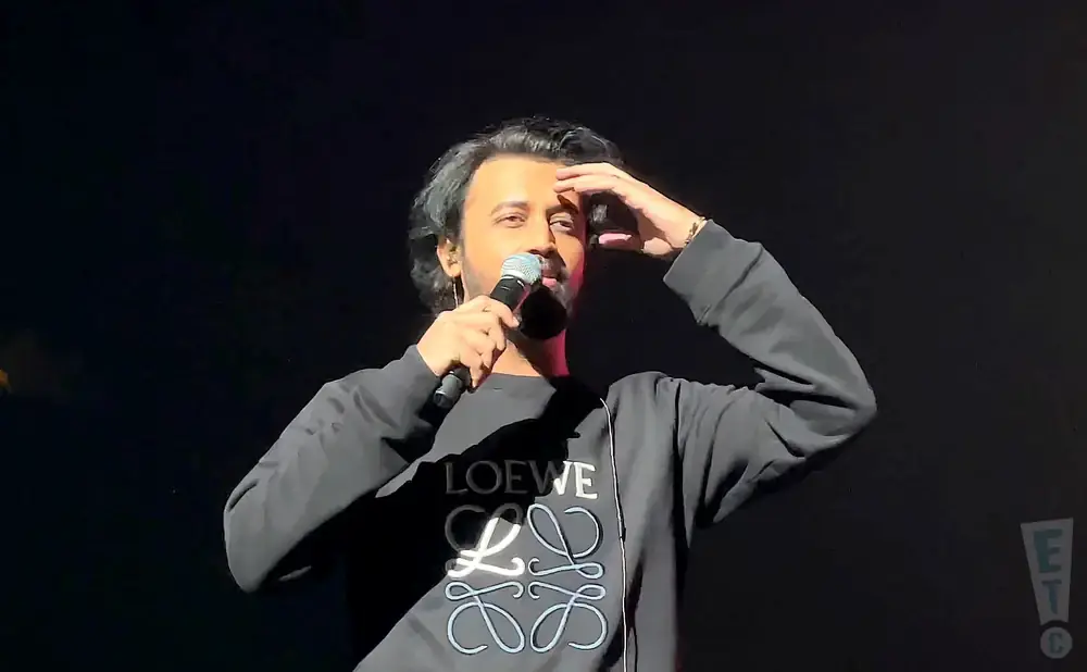 A man on stage holds a microphone and touches his forehead, wearing a black sweatshirt with the 