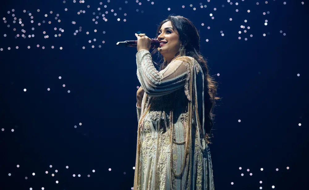 A woman in a sparkling dress sings into a microphone against a dark blue background with bokeh lights.
