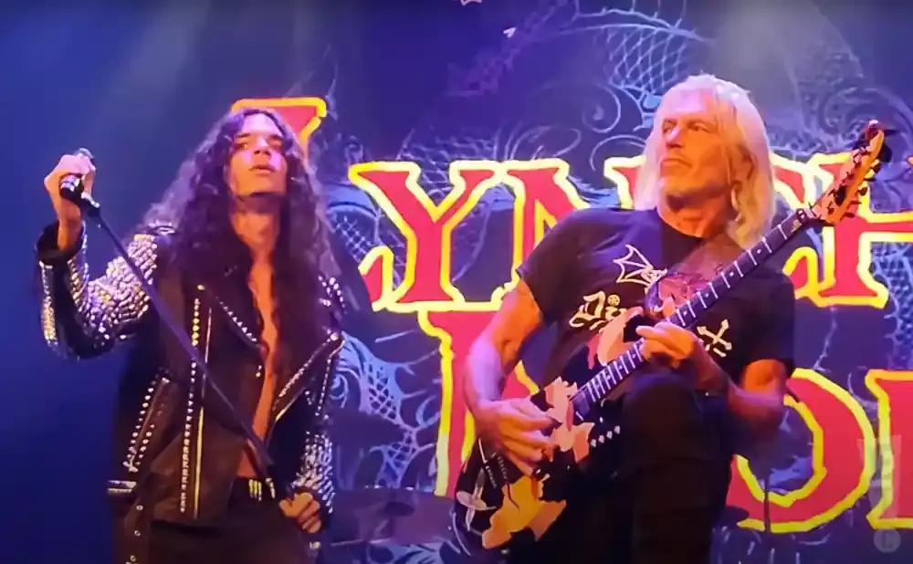 Two male rock musicians perform on stage with a band logo behind them.