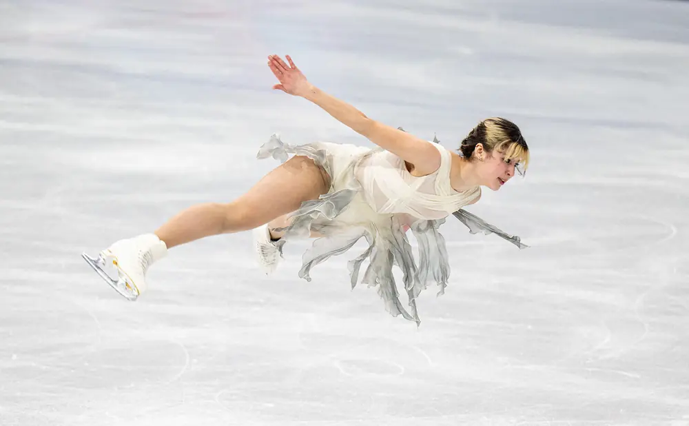 A figure skater in a light-colored dress leaps in the air, her arms outstretched.