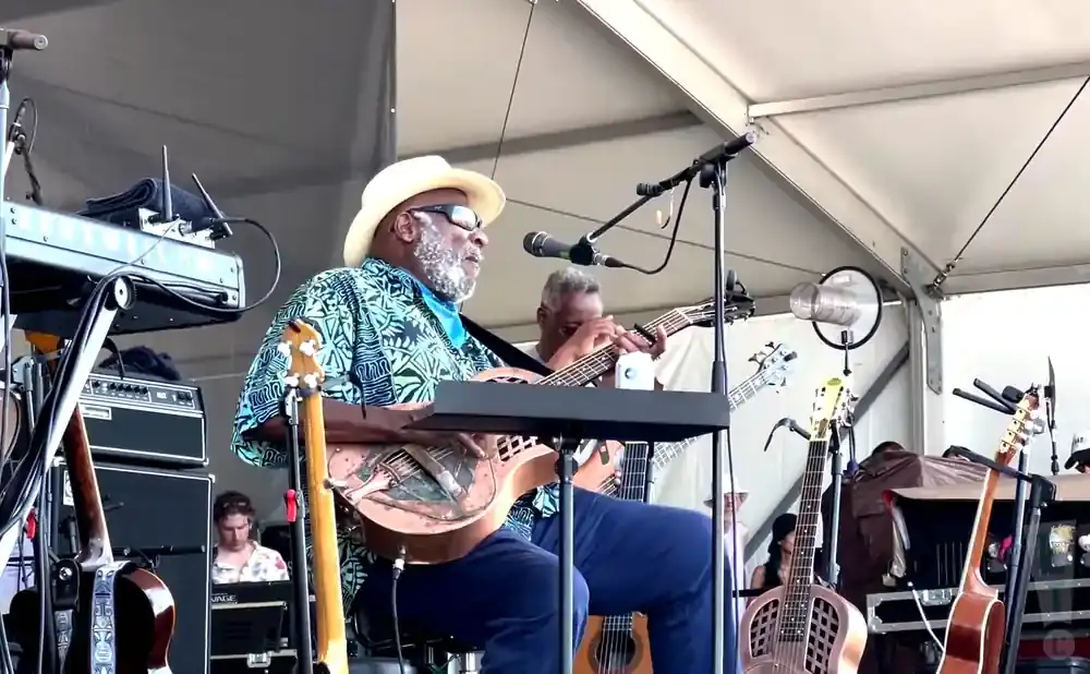 A man in a hat plays a lap steel guitar on stage with other musicians and instruments.