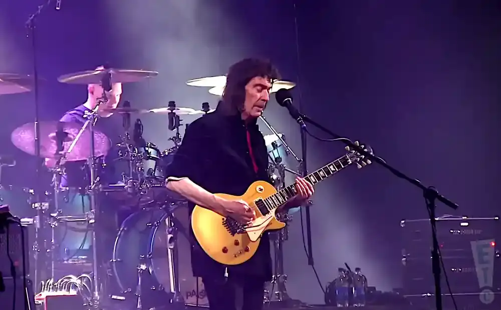 Guitarist Steve Hackett plays a gold electric guitar on stage with a drummer in the background.