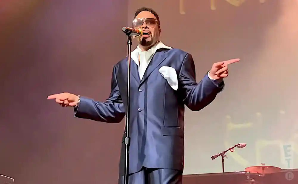 A man in a blue suit and sunglasses sings into a microphone while pointing with both hands.