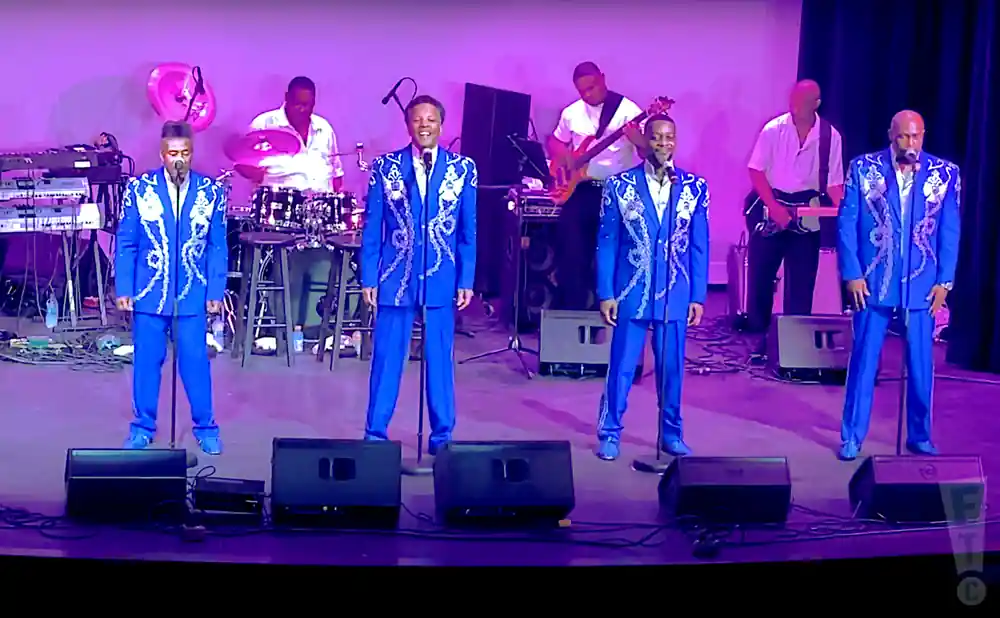 Five men in matching blue suits with silver accents perform on stage, with band members playing drums, bass, and keyboards behind them.