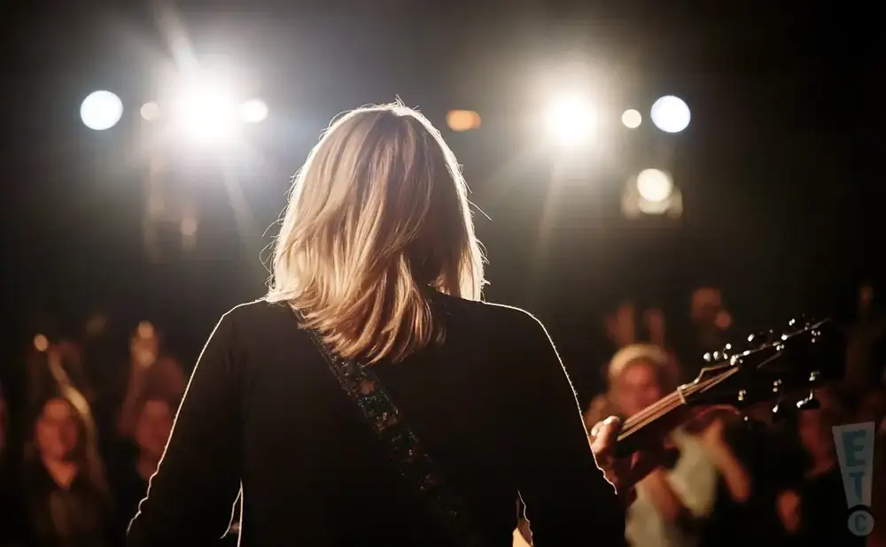 A musician with blonde hair plays guitar on a dimly lit stage with bright spotlights and an audience in the background.