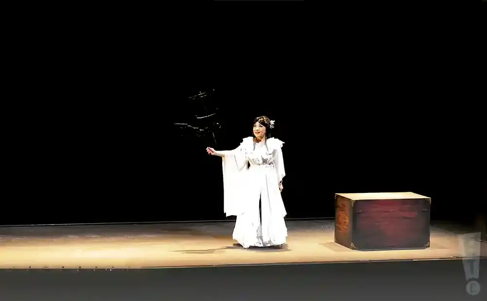 A woman in a white gown stands on a stage, holding a prop. A wooden chest sits to her right.