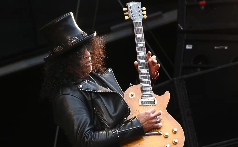 A guitarist with curly hair wearing a top hat and leather jacket plays a Gibson Les Paul guitar.