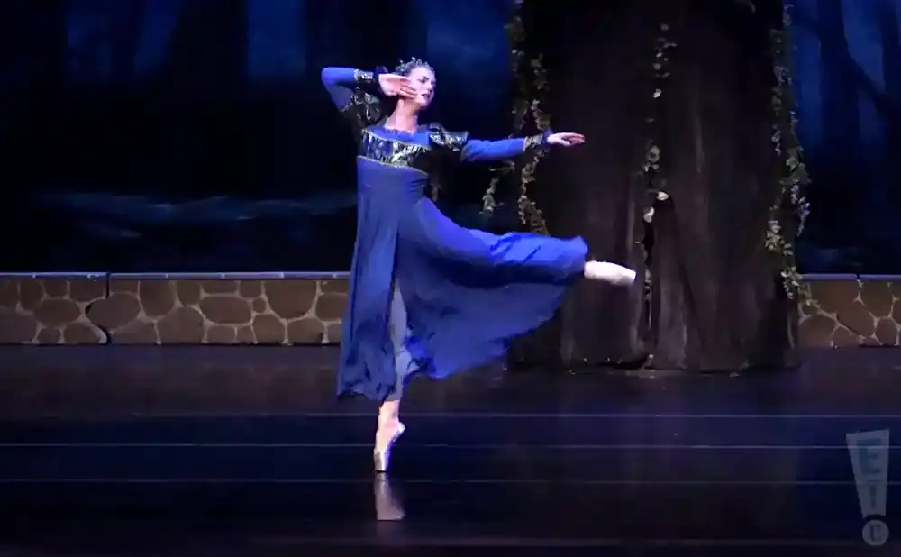 A ballerina in a blue dress and crown performs a graceful pose on stage.