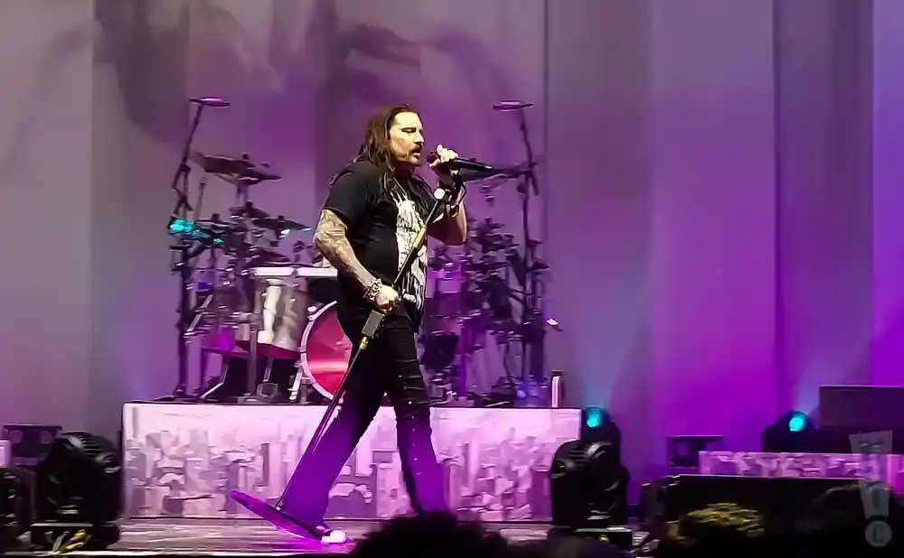 Singer with long dark hair wearing a black t-shirt and black pants, holding a microphone on stage with purple lighting.