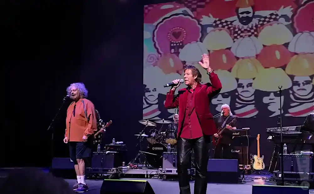 A male singer in a red jacket and leather pants performs on stage with a band behind him.
