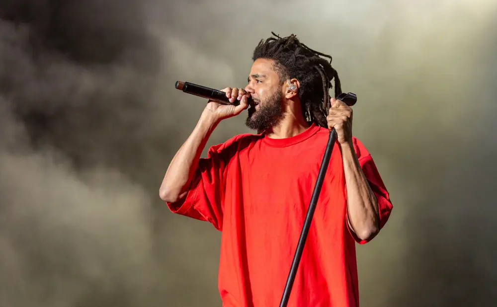 Rapper J. Cole performs on stage, wearing a red shirt and holding a microphone.