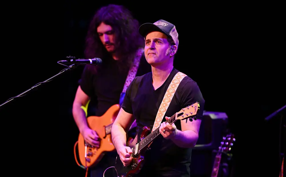 A man in a baseball cap plays a guitar while another man with long hair plays a guitar behind him.