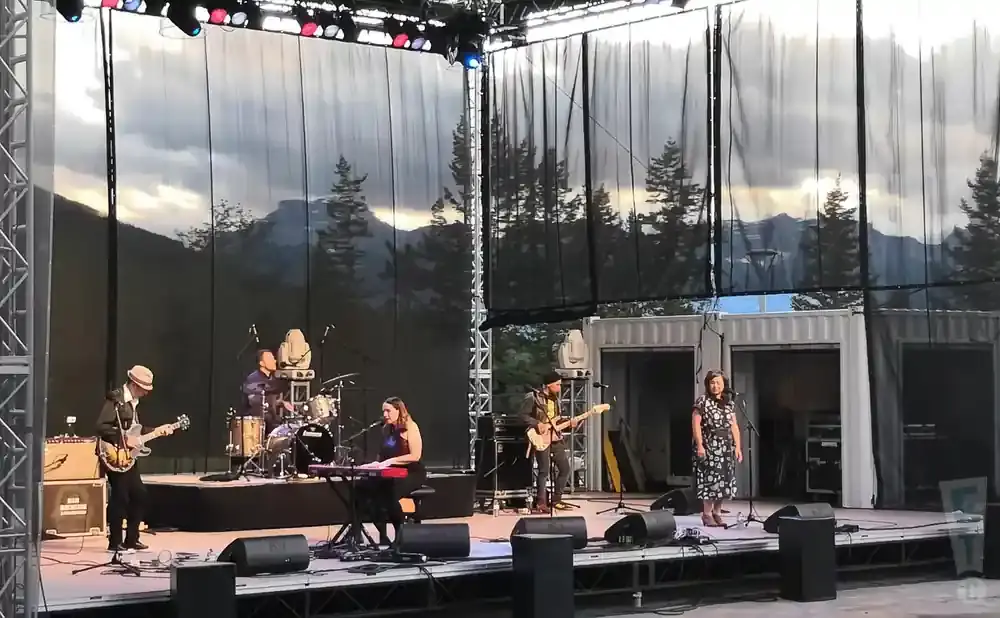 A band performs on an outdoor stage with a mountain and tree backdrop.