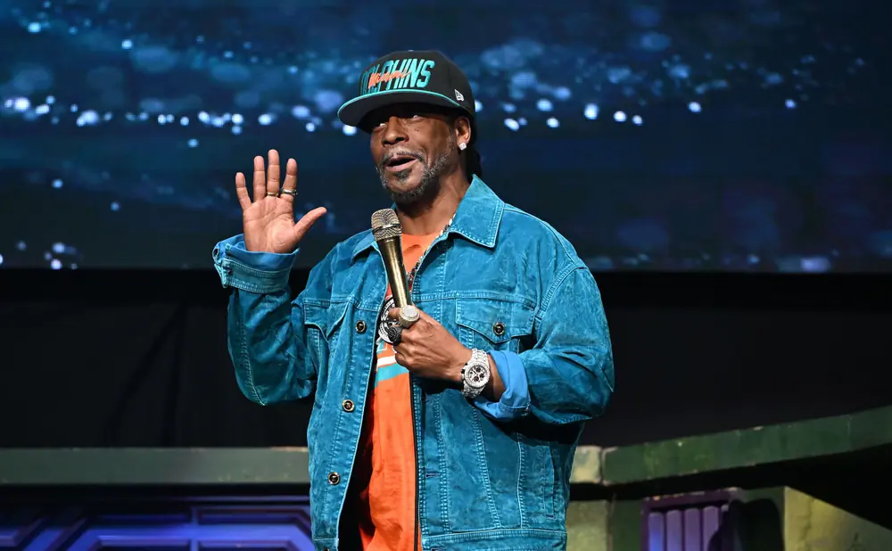 Comedian Katt Williams wearing a teal denim jacket and a Miami Dolphins hat on stage.