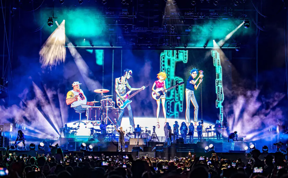 A band performs on a stage with animated characters projected behind them, while a crowd records them on their phones.