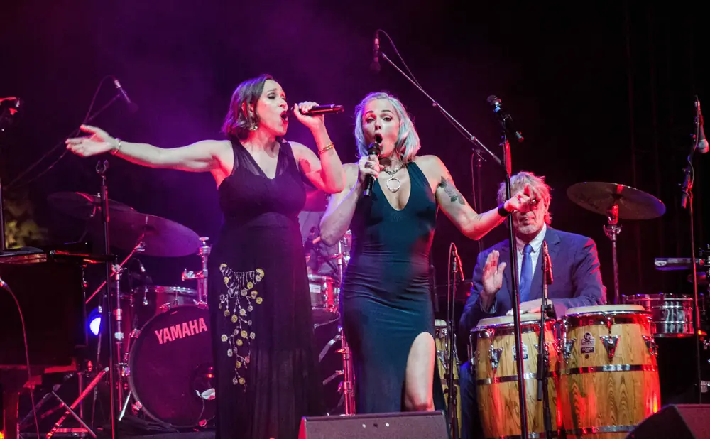 Two women sing into microphones on stage with a band.